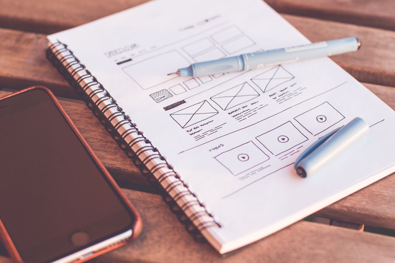Close-up of a notebook showing wireframe sketches and a smartphone resting on a wooden desk.